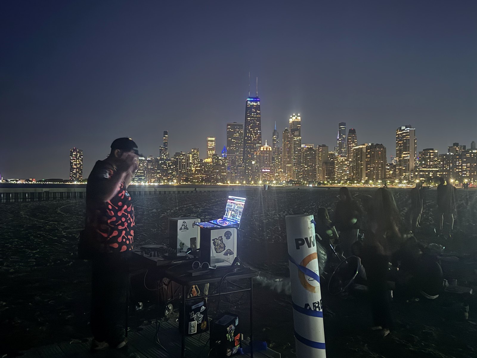DJ DHow performing at a Chicago event with the city skyline visible at night.